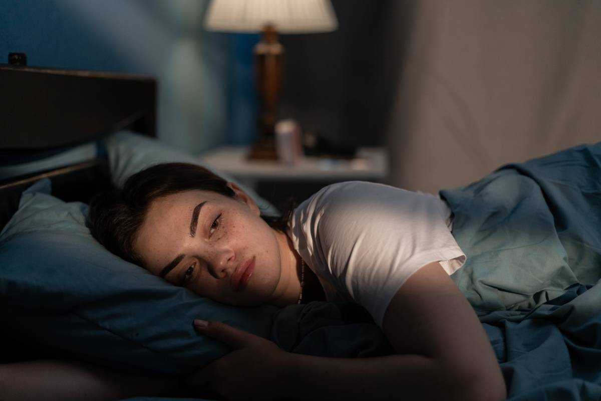 The image shows a clearly sleep deprived woman lying in bed with forlorn expression on her face. It serves to visual represent how sleep deprivation impacts mental health.