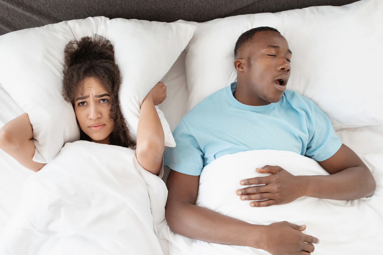 The image depicts a snoring man sleeping with apnea and sleepless woman lying awake in bed next to him. the image represents how snoring affects your relationships.